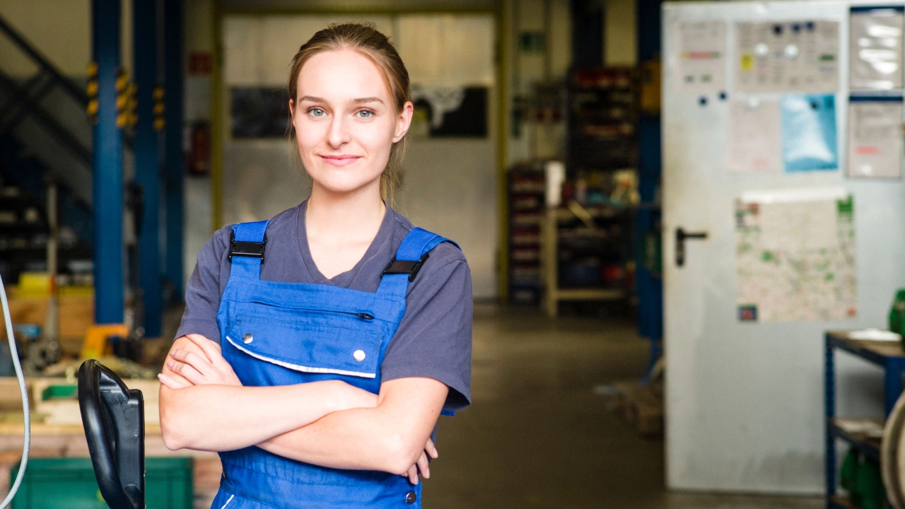 Eine junge Frau im Betrieb in einer blauen Arbeitshose und einem blauen T-Shirt steht mit verschränkten Armen da und schaut freundlich