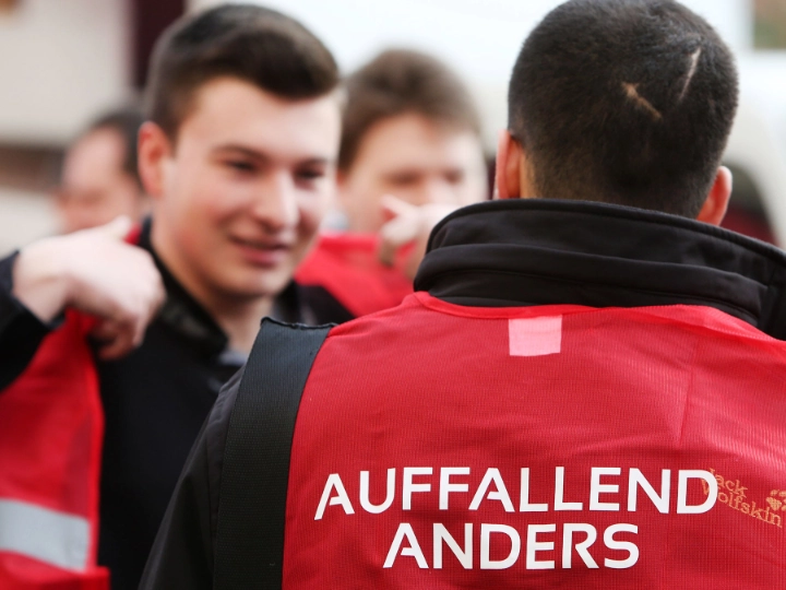 Junger Mann mit roter Weste mit der Aufschrift "Auffallend anders" von hinten.